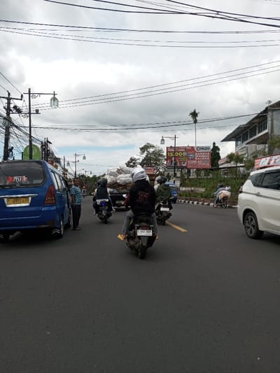 The scene depicts a moderately busy urban street in Taohom, Laos, under an overcast sky with significant cloud cover. The dark asphalt roadway features a yellow solid line, indicating a lane division for traffic moving away from the viewer.
On the far left, a blue minivan, possibly a Nissan Serena, is stopped or parked. Its rear license plate reads "F 3993 ZE". A male individual, wearing a light blue patterned short-sleeved shirt and dark pants, stands next to the driver's side of the minivan, looking towards the right. A dense network of black utility wires crisscrosses overhead, supported by multiple utility poles equipped with decorative streetlights.
In the foreground and central portion of the road, traffic primarily consists of motorcycles moving away from the viewer. The closest motorcycle is a black scooter, ridden by an individual wearing a white full-face helmet, a dark hoodie with white lettering on the back, grey pants, and yellow sandals. The scooter's rear license plate is "F 3611 NBN". Immediately behind this scooter, another rider on a motorcycle, wearing a dark vest and helmet, is visible. Further back, a light blue pickup truck, heavily loaded with large white sacks, is in motion. Several other motorcycles, including one ridden by an individual in a green jacket and helmet, are interspersed within the traffic flow.
On the right side of the road, a white SUV, possibly a Mitsubishi Xpander, is partially visible, moving in the same direction as the other vehicles. The road edge features a black and white striped curb, separating it from a raised area. On this raised area, a large billboard is prominently displayed, featuring text that includes "CEKAS", "15.000", and "BANNER SPANDUK BALIHO". A tall palm tree stands behind the billboard, adjacent to a multi-story building with a grey facade. Further along the right side, another person is visible next to a parked motorcycle.