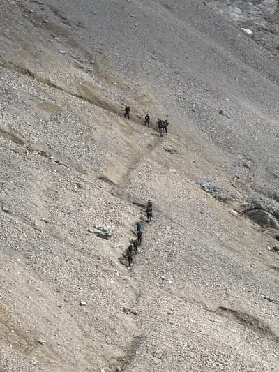 A group of hikers is traversing a steep, gravelly slope in Wildermieming, Austria. The scene is captured from a high vantage point, looking down on the individuals who are spread out along a narrow, worn path. Each hiker is equipped with a backpack and appears to be using trekking poles for stability on the loose terrain. The environment is barren and mountainous, characterized by a vast expanse of small stones and dirt. The sunlight creates strong shadows, accentuating the ruggedness of the landscape. The hikers are moving in a downward direction, implying they are descending the slope or completing a trek.
