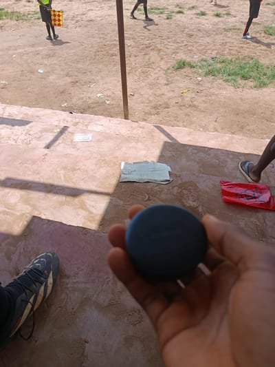 In Ukpor, Nigeria, this outdoor scene captures a dusty, uneven terrain that appears to be a sports field. The ground is a mix of dirt and sparse green grass, with scattered debris suggesting recent activity. In the foreground, a person's hand holds a dark, circular object, possibly a Bluetooth speaker or a similar electronic device. Their left foot, clad in a black and white soccer cleat, rests on the ground, indicating they are likely a participant or spectator at a sporting event.
Further back on the field, another individual is visible, dressed in a bright yellow and black checkered uniform and holding a matching flag, identifying them as a referee or official. Their presence suggests a game is either in progress or has recently concluded. The background shows more of the dusty field and hints of structures or seating areas, including concrete steps in the midground where a crumpled piece of white material and a red plastic bag are placed. The overall atmosphere is that of a casual, community-level sporting event.