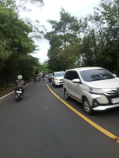 The image shows a road scene with traffic. A motorbike with a rider wearing a helmet is in the left lane. Several cars are in the right lane. The foreground is out of focus, indicating motion. The surrounding environment includes trees on both sides of the road. The sky is overcast. The location is not identifiable as being in Taunggyi, Myanmar, without additional information.