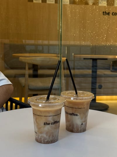 The image displays two clear plastic cups of iced beverages, each containing a black straw, placed side-by-side on a white, circular table. Both cups are branded with "the coffee." text and show distinct layers: a dark brown substance at the bottom, a milky white layer above it, and a lighter brown liquid on top. To the left, a portion of a human forearm, wearing a white shirt, is visible at the table. The background reveals the interior of a coffee shop, viewed through a clear glass partition. This background includes light-colored wooden wall panels, wooden tables with dark bases, and chairs with light upholstery. The "the coffee." branding is also visible on surfaces within the background. The scene is illuminated by warm, internal lighting, creating numerous out-of-focus light reflections on the glass. This establishment is a branch of "the coffee." located in Torreón, Mexico.