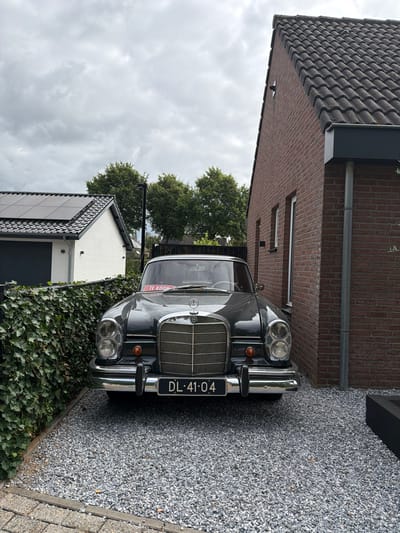 The image captures a vintage dark grey Mercedes-Benz sedan parked on a gravel driveway in front of a modern red-brick house. The car has a license plate reading "DL-41-04" and a red "TE KOOP" (for sale) sign is visible on the windshield. To the left of the car, a white building with a garage door and solar panels on its roof is partially visible, separated by a dense green ivy hedge. The scene is set under an overcast sky, with trees visible in the background. The location is identified as Melick, Netherlands.