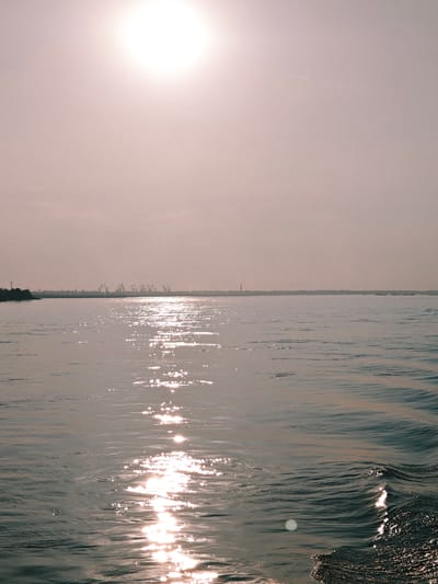 This is a wide-angle, vertical, eye-level shot of a body of water, likely the Angara River, in the city of Lesosibirsk, Russia. The sun is high in the hazy, light-pink sky, creating a bright, diffused light that casts a shimmering reflection on the water's surface. The water itself is calm with gentle ripples and waves, especially in the foreground where a wake is visible. The horizon line is low in the frame, with a distant shoreline visible. Silhouetted against the bright sky are several industrial cranes, suggesting the presence of a port or industrial area along the river. The overall atmosphere is serene and expansive.