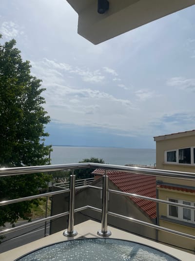 The image shows a view from a balcony overlooking the Adriatic Sea in Dugi Rat, Croatia. The balcony is made of concrete and features a stainless steel railing. A round glass table sits on the balcony, with a view of the sea and surrounding buildings. The sky is a light blue with white clouds. A large green tree is visible to the left of the image. The railings and the table are reflective, showing the surrounding environment. The scene is devoid of people. The building in the image is likely located on the waterfront, providing an unobstructed view of the sea. The scene is bright and sunny, giving a sense of calm and tranquility.