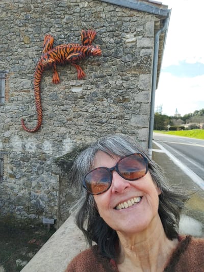 An outdoor daytime photograph captured in Francillon-sur-Roubion, France, depicts a woman in the foreground smiling directly at the camera. She has grey hair, wears dark-framed sunglasses that reflect the surrounding stone and sky, and is dressed in a brown, textured top. Behind her, a large, orange and black striped lizard sculpture is vertically affixed to a rustic stone wall composed of irregular grey and tan stones; its head faces right and its tail curls downwards. To the left of the sculpture, a small window with wooden shutters is visible. The stone building features a grey gutter system along its roofline. To the right of the building, a paved road with a white solid line extends into the distance, flanked by green grass and sparse trees. A distant structure, possibly a water tower or silo, stands on a hill against a partly cloudy blue sky.