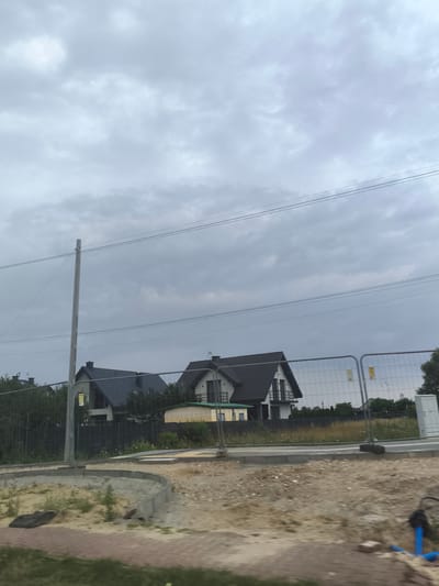 The image depicts a cloudy sky over a residential area in Skrzeszew, Poland. In the foreground, there is a construction site with a curved concrete border, sand, and some debris, including a black mat and blue pipes. A temporary metal fence encloses the construction area. Beyond the fence, two modern houses with dark tiled roofs are visible. The houses have white facades with dark window frames and balconies. A light-colored temporary building or structure is situated between the two houses. Power lines are strung across the upper portion of the image, supported by a tall wooden pole on the left. The overall scene suggests ongoing construction and development in a suburban environment.