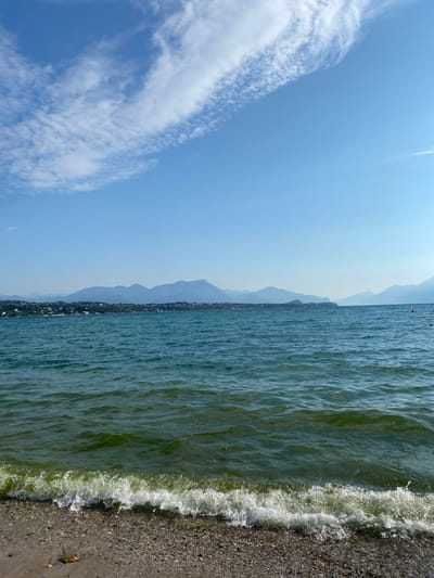 The image depicts a serene lake scene with a pebble beach in the foreground. Gentle waves are rolling onto the shore, with white foam cresting. Beyond the lake, a distant shoreline is visible, lined with buildings and trees, and backed by a range of mountains under a clear blue sky with scattered wispy clouds. The water is a rich green hue, and the sky is a bright, cloudless blue with streaks of white cirrus clouds. The image appears to be taken from a beach in Lonato del Garda, Italy, looking out over Lake Garda towards the mountains. There are no people or specific identifiable objects in the foreground or water, emphasizing the natural beauty of the landscape.