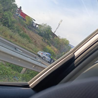 In Dolno Varshilo, Bulgaria, a police car is parked on the side of a road next to a crash site. The crash involves a red truck that has gone off the road and is partially overturned on a grassy embankment. Two police officers, wearing high-visibility vests, are standing next to the police car, seemingly assessing the situation. The police car is white with blue lights and has "ПОЛИЦИЈА" (Police) written on its side in Cyrillic. The environment includes a road with a metal guardrail, green vegetation, and a blue sky with some clouds. The image appears to be taken from inside another vehicle, suggesting a passing observation of the scene.