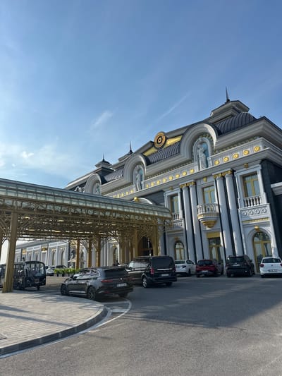 The image displays the exterior of a large, ornate building in Kapitan Andreevo, Bulgaria, under a clear blue sky with scattered cirrus clouds. The building's architecture is characterized by classical and Baroque revival elements, featuring a predominantly white and dark grey facade with extensive gold detailing on cornices, window frames, balustrades, and column capitals. The roofline includes multiple dark-tiled domes and pointed finials. Sculptural figures are visible within arched recesses on the upper levels. A prominent, gold-colored metal and glass canopy extends over a paved driveway, serving as a portico.
Several vehicles are present on the asphalt driveway and parking areas. A black Renault Talisman Grandtour station wagon, bearing Romanian license plate B 123 CAH, is positioned on the driveway, turning. A small, open-sided black utility vehicle is parked near the base of the canopy. Underneath the canopy, a black Mercedes-Benz Vito van with Bulgarian license plate CB 1067 CE is parked. Further along the building's facade, other parked vehicles include a white sedan, a red hatchback, a dark Jeep Renegade with Bulgarian license plate CM 1912 MX, and a white Volkswagen Polo hatchback with license plate 22 ML 747. The ground consists of asphalt for vehicular traffic and lighter grey paver stones for pedestrian pathways and curbs. The scene is illuminated by direct sunlight, casting distinct shadows. Vague human figures are discernible in the distance near the building's entrance.