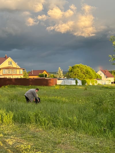 A medium, landscape-oriented shot shows a man kneeling in a grassy field in Vysokinichi, Russia. Storm clouds fill the left sky, while residential houses line the horizon.
The man, dressed in a grey jacket and black pants, is kneeling in the middle-right of the frame. He holds something dark in his hands.
Behind him, a brown fence separates the field from a row of houses. The houses are varied in color and style, with one being yellow and another having a red roof.
The field is covered in tall green grass, with patches of lighter and darker shades. The sky is filled with dramatic storm clouds on the left, contrasting with the sunlight on the right. Some trees can be seen in the background, along with a few other buildings.