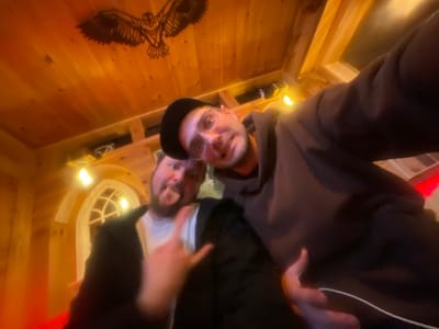 A low-angle, slightly tilted selfie captures two men in a warmly lit, rustic indoor setting. The man in the foreground, wearing a dark cap and glasses, holds the camera, his face partially visible with a slight smile. Behind and to his left, the second man, sporting a beard, is making a playful expression by sticking out his tongue and forming a "rock on" hand gesture. He wears a white shirt under a dark jacket.

The background features extensive wooden paneling on the walls and ceiling, contributing to a cozy and inviting atmosphere. A prominent, intricately carved wooden decoration of an owl or bird in flight is affixed to the wooden ceiling directly above the subjects. Warm-toned artificial lights illuminate the space, suggesting an evening or nighttime setting. To the left, a distinctive window with a Gothic-style arched frame and cross-pane design adds to the unique architectural character of the room. The rustic, lodge-like interior, with its natural wood elements and unique decor, evokes a setting that could be found in a mountainous or rural region, such as Moračko Trebaljevo, Montenegro. No visible text is present.