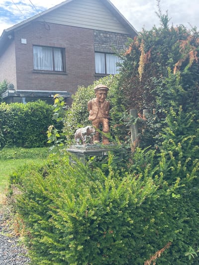 The image displays a garden scene in front of a brick house. A wooden sculpture of an elderly man wearing a cap and jacket stands next to a sculpture of a dog. The sculptures are placed on a pedestal in a garden surrounded by dense green bushes and a lawn. The house, partially visible in the background, has brick walls and a tiled roof. A window with white curtains is visible on the side of the house. The garden appears to be well-maintained, with various types of greenery. The weather seems overcast, with a cloudy sky. This scene is located in the city of Diepenbeek, Belgium.