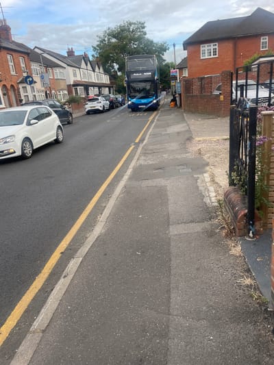 The image is a street scene in Reading, United Kingdom. A blue double-decker bus is traveling down a road lined with houses and parked cars. There is a pavement on the right side of the road with a black metal gate and a brick wall. The sky is cloudy. A white car is parked on the left side of the road. The road has yellow double lines painted on it.
