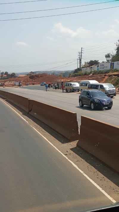 The scene depicts a daylight view of a multi-lane asphalt road in Gitaru ward, Kenya. In the foreground, a dark asphalt road with a solid white edge line runs alongside a series of weathered, brown concrete road barriers exhibiting visible cracks. In the midground, beyond these barriers, a dark blue/black Volkswagen Tiguan SUV (license plate KCE 245A) travels leftward. Further along the opposite side of the road, several white passenger vans, including one with a license plate starting 'KBU', are stationary or parked at the roadside. Approximately 10-15 individuals are gathered near these vans, some appearing to engage in roadside vending or interactions, while two individuals walk away from the group. The background features extensive areas of exposed red earth, indicating construction or excavation, with scattered residential buildings visible on distant hills. Utility poles with multiple power lines traverse the upper frame, and a billboard partially displays text including "Vinyl Matt". The sky is light blue with sparse white clouds.