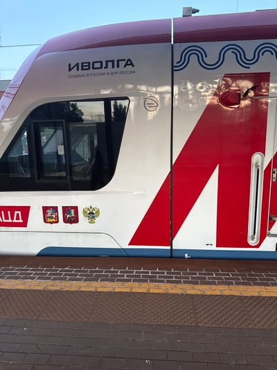 The image shows the side of a white and red train with the word "ИВОЛГА" (Ivolga) written in black text, along with the phrase "СОЗДАНО В РОССИИ И ДЛЯ РОССИИ" (Made in Russia and for Russia). There are also three emblems on the train's side: two red shields with knights on horseback and a golden double-headed eagle. The train is parked at a station with a brick platform and yellow tactile paving. The environment is outdoors, with a clear sky visible. There are no people visible in the image. The specific location in Mamonovo, Russia is not indicated within the image itself.