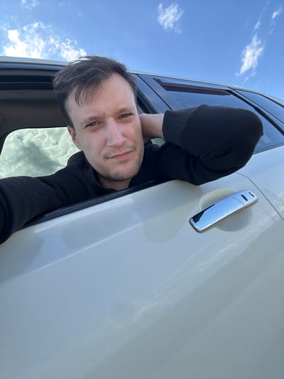 A man is captured in a close-up, outdoor selfie taken from inside a vehicle. He is looking directly at the camera with a relaxed expression. His left arm is resting on the car door frame, with his hand behind his head. He is wearing a black hoodie. The sky, with scattered clouds, is visible in the background. The exterior of the car, which is white, is also visible, including the chrome door handle. There is no specific indication of the city of Abrykosivka, Ukraine.