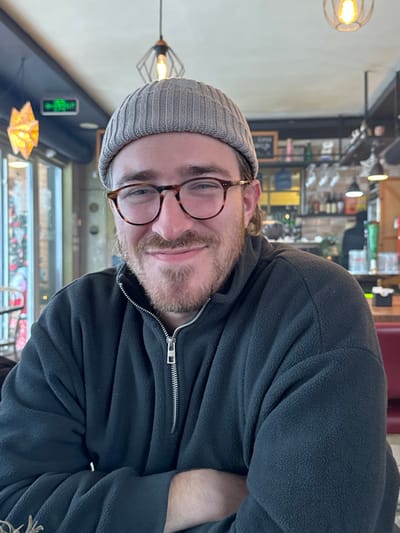 The scene depicts an indoor establishment, likely a cafe or bar, located in Raska Municipality, Serbia. A single adult male is positioned centrally in the foreground, facing directly towards the viewer with a closed-mouth smile. He appears to be in his late 20s to early 30s, with light skin, a trimmed light-brown beard and mustache, and short light-brown hair visible beneath a light grey knitted beanie. He is wearing tortoise-shell framed eyeglasses and a dark grey fleece pullover with a silver half-zipper. His arms are not fully visible but appear to be crossed or resting.

The background to the left features a large window admitting natural light, through which blurred outdoor elements, including a red decorative object or festive lighting, are discernible. An illuminated green rectangular sign with characters resembling Asian script is mounted on an interior wall near the window frame. A decorative, star-shaped light fixture with a warm glow hangs near this window.

The background to the right reveals a bar or counter area. Shelves display various bottles and inverted wine glasses. A section of red upholstered seating is visible behind the subject's right shoulder. Overhead, two pendant light fixtures with exposed Edison-style bulbs, one enclosed in a black geometric metal frame, illuminate the space. A dark board, possibly a menu, is partially visible above the bar. The ceiling is light-colored.