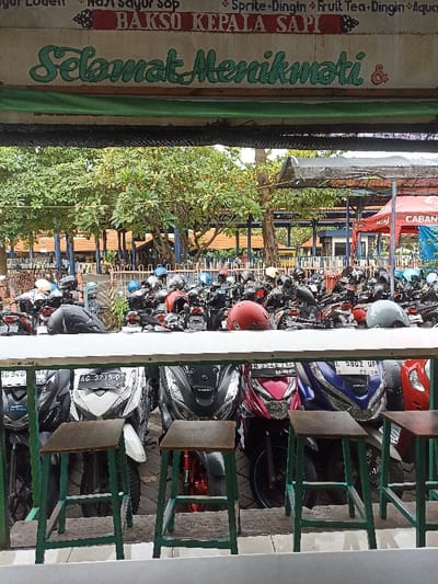 The scene is captured from inside a food stall with the sign "Bakso Kepala Sapi," which translates to "Ox Head Meatballs." The sign also says "Selamat Menikmati" meaning "Enjoy Your Meal". The image shows a view of the street from inside the stall. There are numerous parked motorcycles behind a low wooden fence. In the foreground, there are several motorcycles parked on the street. Four of them are positioned close together in a row, with the front wheels facing the viewer, while the remaining motorcycles are parked further back in the image. The image is taken from a low angle, showing the motorcycles and the street below. The area behind the motorcycles is a parking lot with trees and a sign reading "Caban." The location of the stall is in Kecamatan Waru, Surabaya, Indonesia. It is a sunny day. The motorcycles are parked on a paved street. The environment is a busy urban area, with trees and buildings in the background. There are no people visible in the image.
