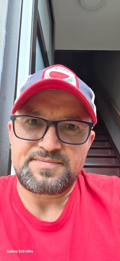 The image shows a close-up, sideways selfie of a person wearing glasses and a baseball cap. The person has a beard and is wearing a red t-shirt. The baseball cap is blue and white with a red brim and a circular patch featuring a slice of watermelon. The background appears to be an indoor setting with dark paneling and a window. The image has a watermark that reads "Galaxy S26 Ultra". The overall impression is a casual, personal photograph.
