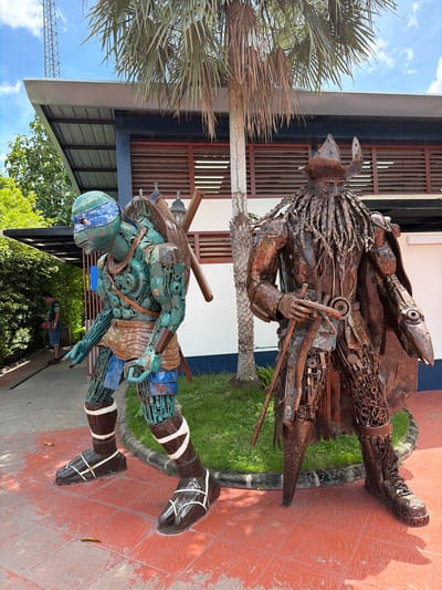 In the city of Tha Ruea, Thailand, two statues are displayed outdoors. The statue on the left depicts a Teenage Mutant Ninja Turtle character, likely Leonardo, as evidenced by the blue mask and the katanas (though not clearly visible in this pose). This statue is constructed from various metal components, including what appears to be rusted metal and blue painted metal. The statue on the right is made entirely of rusted metal and depicts a figure wearing a helmet and a cape, with a sword in its hand. The statues are placed on a reddish-orange tiled ground in front of a building with a metal roof and wooden louvers. Green grass is visible behind the statues, and palm trees are present in the background. A person wearing a t-shirt and shorts can be seen in the background to the left, near some green foliage. The sky is blue with some clouds.