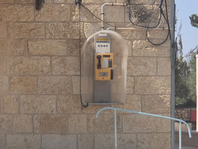 An exterior shot captures a weathered, light brown stone wall with visible mortar. Mounted on the wall is a clear plastic enclosure housing a yellow public payphone. The phone has a dark grey handset, a keypad, and a slot for coins or cards. Above the phone, a sign displays the number "5330509" and other text in Hebrew and English. Electrical wires, both black and white, are routed along the wall and into the enclosure. A light blue metal railing, curved at one end, extends from the bottom right of the frame upwards towards the left. In the background, trees and a blue sky are visible. The setting appears to be the historic village of Abu Gosh, Israel, a location known for its ancient architecture and cultural significance.