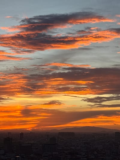 This image depicts a sunset over the city of Khan Mean Chey, Cambodia. The sky is filled with dramatic clouds painted in vibrant hues of orange and red, transitioning to softer yellows and blues towards the top. Below the colorful sky, a sprawling cityscape is visible in silhouette, characterized by numerous buildings of varying heights, including taller structures and what appear to be residential areas with lower-rise buildings. In the distance, a range of mountains forms a hazy silhouette against the horizon. The overall scene conveys the beauty of a sunset in an urban environment with a natural backdrop.
