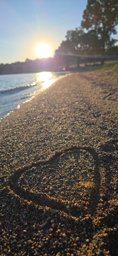 A close-up, low-angle shot of a pebble-strewn beach on a sunny day. The sun, low in the sky, casts a warm glow on the scene, with its reflection shimmering on the gentle waves of a lake. In the foreground, a heart shape is drawn in the pebbles. The background shows a line of trees and a blurred horizon. The scene is peaceful and evokes a sense of romance. This image was taken in Issyk-Kul, Kyrgyzstan.