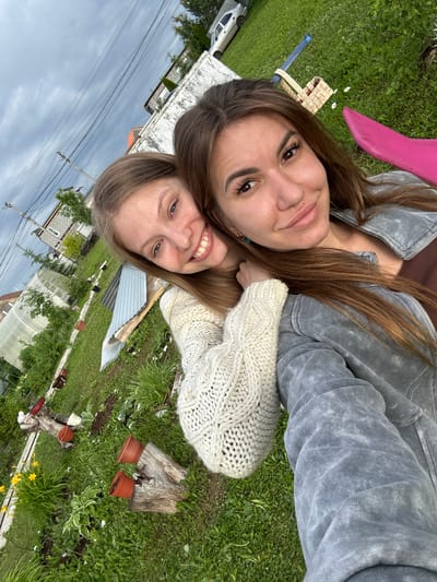 This image captures two females posing for a tilted selfie in an outdoor environment within Bol'shaya Mos', Russia. The sky is overcast with dense grey clouds.

The female on the left, appearing in her late teens or early twenties, has long light brown hair and is smiling broadly, exposing her teeth. She is wearing a cream-colored, cable-knit long-sleeved sweater. Her left arm is positioned around the shoulders of the female on the right.

The female on the right, appearing in her twenties or early thirties, has long brown hair and a more subdued smile. She is wearing a grey, textured or acid-washed zippered jacket over a brown top, with a small teal earring visible on her left earlobe. Her right arm is bent, with her hand resting on the left arm of the other female.

The background consists of a green grassy area resembling a garden or yard. Visible elements include several terracotta plant pots, some empty and some containing small plants, placed on wooden stumps. Yellow daylilies and other green foliage are present. A large, translucent white greenhouse structure and a stack of corrugated metal sheets are visible in the midground on the left. Further back, residential or utility buildings with dark roofs are seen. Multiple utility poles with power lines extend horizontally across the upper left portion of the frame. On the right side of the background, a woven wicker basket with a blue handle lies on the grass near small white flowers. A silver sedan is parked in the distance, and a bright pink object, possibly a plastic item, is partially visible in the upper right corner.