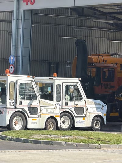 The image shows two airport vehicles parked next to each other in an outdoor setting. The vehicle in the foreground is a white tow tractor, with "welcome AIRPORT SERVICES" printed on its side. The number "1122" is visible on the lower part of the tractor. The vehicle behind it is orange and appears to be a larger airport vehicle, possibly a loader or a snow plow, with the letters "PWS" visible on its side. Both vehicles are parked on a paved surface next to a grassy area and a concrete curb. The scene appears to be during the daytime, with sunlight casting shadows. In the background, there is a building with a corrugated metal roof, suggesting an airport hangar or service area. A blue circular sign with a white arrow pointing left and a red circular sign with the number "5" are visible on the left side of the image, indicating directional or informational signage within the airport environment. The overall impression is one of airport operations and infrastructure.