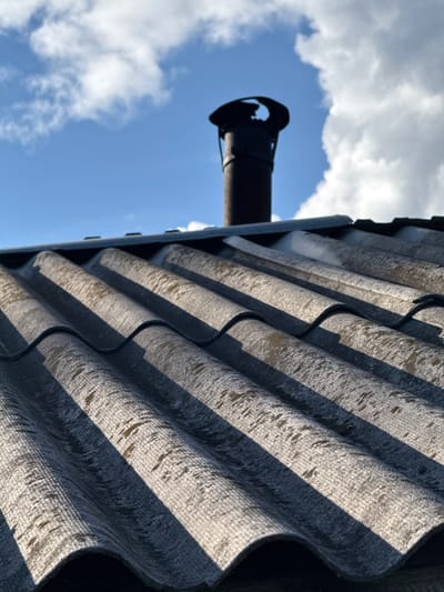 The image shows a section of corrugated asbestos cement roofing material, oriented diagonally from the upper right to the lower left of the frame. The material exhibits a repeating wave pattern, with alternating bands of light and shadow due to the sun's angle. A black metal chimney pipe extends vertically from behind the roofing, positioned on the left side of the image and silhouetted against a blue sky with scattered white clouds. The location is unidentified; there is no identifiable information to confirm this photo was taken in Medvedka, Russia.
