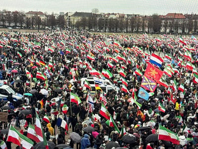 A wide-angle aerial view captures a massive outdoor gathering of thousands of individuals assembled in a large open space within the city of Vellmar, Germany. The scene is overcast, with numerous participants holding open umbrellas in various colors (primarily black) against potential rain or drizzle.
The crowd is densely packed and extends into the far background. The predominant objects are flags, with a significant majority being the historical Iranian imperial flag (featuring green, white, and red horizontal stripes with a golden lion and sun emblem at its center). Other flags present include plain green, white, and red tricolor flags, the national flag of the Netherlands (red, white, and blue horizontal stripes), and several distinct banners with unique symbols (e.g., a golden starburst on a red field, a purple motif on a yellow field, and a light blue flag with a central symbol). These flags are held aloft by individuals throughout the assembly.
In the foreground on the left, a white and blue German police vehicle (marked "Polizei") is partially visible. The background reveals a line of multi-story buildings with traditional European architecture and bare deciduous trees, indicative of an urban or suburban setting during a cooler season. Participants are largely stationary, collectively forming a large-scale demonstration or protest. No specific landmarks within Vellmar are discernible to pinpoint the exact location.