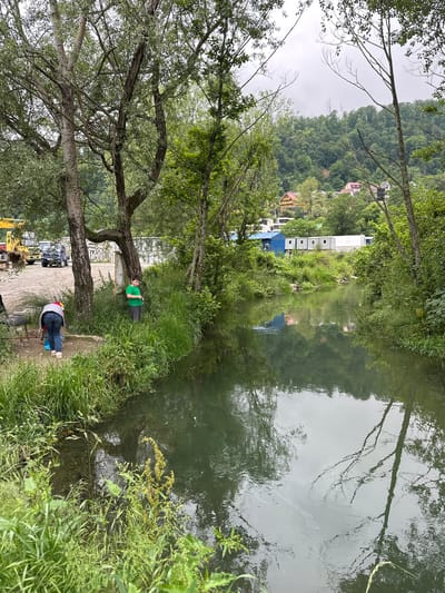 The image captures a tranquil scene along a waterway, possibly a river or canal, in what is presumed to be Bolshoy Sochi, Russia, based on the presence of characteristic architecture in the background. The viewpoint appears to be from the bank, where a partially visible yellow excavator and construction materials hint at ongoing development or maintenance.

Two individuals are prominent. One, wearing a green shirt, reclines against a tree near the water's edge, seemingly relaxing or resting. The other person, sporting a red shirt and a grey hat, is crouched near the bank, possibly fishing, with a fishing rod and chair visible near them.

The waterway reflects the surrounding vegetation and some distant buildings, suggesting relatively calm conditions. Trees line the banks, with their branches extending towards the water, and lush green foliage is abundant. The background features a hillside dotted with buildings in various colors, a common sight in coastal Russian towns like Bolshoy Sochi. The sky appears overcast, casting a soft, diffused light across the scene.