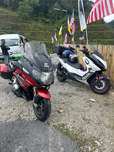 A bright, overcast day features two motorcycles parked on a gravel surface next to a bamboo fence. In the foreground, a red and black BMW motorcycle is parked facing left. The motorcycle has a license plate that reads "VRF 8104". Behind it and to the right is a white and black scooter with purple accents. This scooter has the number "0" in a black square on its side. Both vehicles have luggage racks and helmets are placed on the seats of the scooter. In the background, several Malaysian flags are flying on poles, and a white van is partially visible to the left. A man in a black jacket and jeans stands with his arms crossed next to the van, and another man in a patterned shirt is near the van's doorway. The environment appears to be a roadside area in a hilly, green region, possibly Kampung Kerawat, Malaysia, characterized by terraced hillsides covered in lush vegetation.