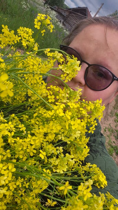 A person wearing sunglasses is holding a large bouquet of yellow flowers in front of their face, obscuring most of their features. The background shows a rural scene with a wooden building, tall grass, and a dirt path. The image appears to be taken outdoors in a natural setting.