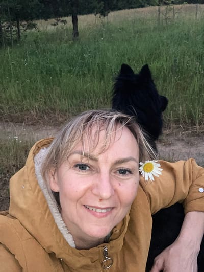 A close-up selfie shows a woman with blonde hair and a daisy tucked behind her ear. She is wearing a mustard-colored jacket with a white, fuzzy lining. A black dog is visible behind her. The background consists of a grassy field with trees and a dirt path. The scene appears to be in an outdoor, rural setting, possibly the vicinity of Міханавіцкі сельскі Савет, Belarus.