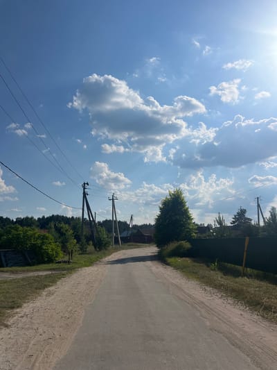 The image shows a dirt road leading through a rural area with a clear blue sky filled with white clouds and the sun shining brightly. Several utility poles with overhead wires are visible along the side of the road. Dense green trees line the background, with glimpses of houses and a dark green fence. The road surface appears to be a mix of asphalt and sand, with tire tracks visible on the sandy portion. The overall impression is a sunny day in a tranquil, possibly suburban or village setting in Podlesny, Russia. There are no people or animals in the frame, and no discernible actions or interactions are taking place.