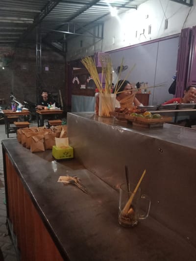 This image depicts a casual dining establishment, likely a warung or small restaurant, located in Cangkring, Indonesia, captured in low-light conditions. The scene primarily focuses on a service counter in the foreground and patrons seated at tables in the midground and background. In the foreground, a metallic counter extends diagonally from the lower-left to the right. On the left side of the counter, a stack of approximately twelve small, brown paper bags, folded and sealed, are neatly arranged. Adjacent to these bags is a lime green tissue box with white text. Further right, a clear glass jar contains numerous thin, light-colored skewers. Behind the jar, parts of a clear plastic bottle and a red condiment bottle are visible. A wooden tray containing several pieces of grilled food, possibly chicken or other meat, and four green limes is positioned on the counter, slightly to the right of the skewers. In the bottom right corner of the counter, a clear glass mug with a handle holds a brown spoon and a yellow stirrer. Near the center of the counter, a crumpled piece of paper or tissue lies beside a pair of small metal tongs. In the midground, three individuals are present. To the left, a young male with short dark hair, wearing a dark t-shirt, is seated at a simple wooden table with a wooden stool. He is looking upwards and slightly to the right. A small white container and other indistinct items are on his table. Behind him, a motorbike is partially visible. Centrally, behind the service counter, a person with long dark hair is seated, looking down with a slight smile, seemingly engaged with the items on the counter. To the right, another male with short dark hair, wearing a red t-shirt, is seated at a different wooden table, laughing with an open mouth and looking towards the right side of the frame. The background reveals the establishment's structure. On the left, an exposed brick wall is visible, with a metal support beam extending vertically. The ceiling features a corrugated metal roof with visible metal trusses. On the right side of the background, a light grey wall with several small, vertical rectangular cutouts is present. Further right, a purple, possibly corrugated metal or plastic, structure resembling a gate or partition is visible. Above this, red and white triangular banners are strung across. The overall lighting is dim, suggesting evening hours,