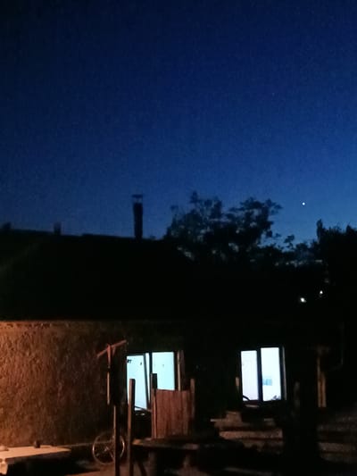 This is a nighttime, low-light photograph taken in Leușeni, Moldova. The scene depicts a dark, rural residential property with a building that appears to be a house or a barn. The sky is a deep, twilight blue, with a single bright star or planet visible in the upper right quadrant. Silhouetted against the sky are trees and what appear to be two chimneys on the roof of the structure. The building's walls are textured and dark, with two lit windows on the ground floor. The left window is a large, single pane, while the right window is divided into two panes. Outside the building, there are various objects, including what looks like a bicycle leaning against a post, a wooden gate or door, and some wooden structures that could be part of a shed or porch. The overall impression is one of quiet and darkness, with the illuminated windows providing the only significant light source on the property.