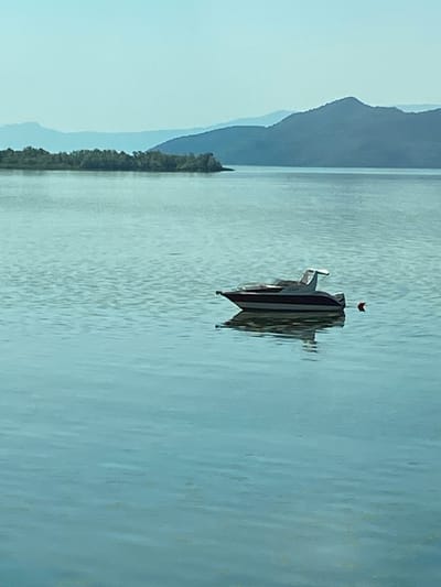 An aerial drone shot captures a tranquil scene in Kruševica, Montenegro. A sleek, dark-hulled motorboat with a white canopy is anchored in calm, turquoise waters. The boat is positioned to the right of the frame, its reflection subtly visible on the water's surface. A small, red buoy floats nearby. In the distance, a lush, green island dotted with trees lines the horizon, transitioning into layers of distant, mist-shrouded mountains. The sky above is a clear, pale blue, creating a serene and expansive atmosphere.