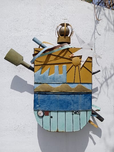 An outdoor mixed-media relief sculpture is affixed to a textured white stucco wall in San Joaquín, Venezuela. The vertically oriented artwork, appearing roughly shield-shaped with a rounded bottom, is constructed from painted metal elements and measures approximately 1.5 times taller than wide.
The sculpture's upper section displays a gold-yellow background with stylized light blue industrial or city structures, a circular yellow sun disc, and a white multi-pointed star. Above these, two wavy gold-tan lines suggest mountains. A distinct dark blue horizontal band separates the upper and lower halves. The lower section comprises vertical light blue-green metal strips, forming a rounded base with a circular metallic element and a small reddish-brown square on the lower-left.
Protruding from the top center is a gold-colored spherical object within an open cage-like structure, topped by a small cross. A green paddle-shaped object extends horizontally from the upper left, alongside an angled light blue cylindrical object. Multicolored metallic strips wrap around the upper body. Dark, irregular objects protrude from the lower right. Strong directional sunlight casts defined shadows to the left and bottom right of the artwork. Sparse dark vegetation is visible against the wall and bright sky in the upper right background.