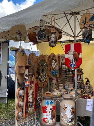The image displays an outdoor market stall in Abondance, France, identified by the distinctive stone spire of the Abondance Abbey Church visible in the background under a partially cloudy sky. The stall operates under a white canopy/umbrella and is densely packed with handcrafted alpine-themed merchandise.
The foreground is dominated by display grids holding various wooden, ceramic, and metal objects. Prominent items include:
* Numerous wooden plaques and wall hangings of various shapes (rectangular, oval, irregular), many featuring painted portraits of brown and white cows, bulls, chamois, and marmots, often surrounded by white edelweiss and blue gentian flowers. Some wooden pieces are functional, such as oval clocks and a small shelving unit.
* Several painted metal cowbells, hanging from leather straps or displayed on racks, also adorned with cow imagery and flowers.
* Two large, painted metal milk cans standing on the ground, each featuring detailed cow portraits and alpine flowers. One milk can also incorporates a red and white shield design, possibly representing a Swiss or Savoyard canton, with a white cross.
* A red, shield-shaped wall hanging with a prominent white cross and a cow portrait, similar to the Swiss flag motif, is attached to a pole.
* An assortment of ceramic items, including pitchers, teapots, and mugs, decorated with rustic alpine scenes and floral patterns, displayed on shelves or tables.
* Small wooden figures and other decorative objects are interspersed among the larger items.
A dark blue vehicle is partially visible on the left side in the middle ground, parked behind the stall. The ground beneath the stall appears to be covered in green grass or dirt. No people are directly visible in the immediate foreground, but the setup indicates an active commercial environment.