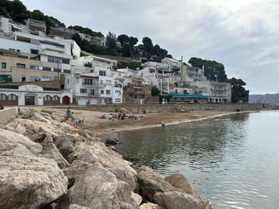 The image presents a coastal view in L'estartit, Spain, featuring a beach and a hillside urban development under an overcast sky. In the immediate foreground, large, textured grey-brown rocks form a low barrier or natural shoreline. The adjacent water body is dark, exhibiting calm ripples, and extends to a distant, hazy coastal landmass on the right.

A narrow strip of light brown sand constitutes the beach. Approximately ten individuals are visible. On the left side of the beach, two adults and two children are standing near the rocky area. Further to the right, a group of about six people are distributed across the sand, some lying down, one beneath a small, light-colored beach umbrella. One individual is partially submerged in the shallow water, bending forward.

The background is characterized by a steep, tree-covered hillside densely populated with white, multi-story buildings. These structures, appearing to be residential or hotel properties, are terraced up the slope, many featuring balconies. A building on the lower left of the hillside is identified as "Bar-Restaurant Bella-Blava" by its signage, featuring arched window or door frames. Dark green, bushy trees are integrated among the buildings and cover the higher sections of the hill. The sky is uniformly cloudy.