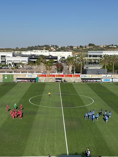 This is an aerial view of a soccer field in Ness Ziona, Israel. A team in red uniforms is positioned at one end of the field, while a team in blue uniforms is at the opposite end. A lone figure in a yellow shirt stands near the center circle of the field. Advertisements for "Winner," "Mega Sport," and "Cargo" are visible along the sidelines. Buildings and palm trees line the perimeter of the field. A blue sky is visible in the background.