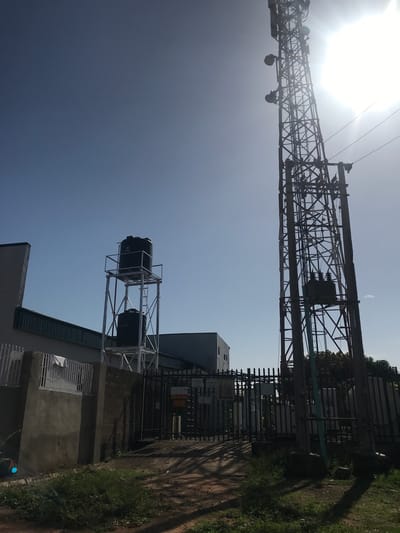 An outdoor, brightly lit image depicts a towering metal lattice structure, likely an electricity transmission or telecommunications tower, under a clear, bright blue sky with a prominent sun causing glare in the upper left. The tower is equipped with various components, including power lines, insulators, and a transformer visible in the mid-section, along with what appear to be dish antennas near its top.
Below and to the right of the tower, a metal fence with spiked tops encloses an area, possibly a substation or industrial facility. Behind the fence, several buildings are visible, including a multi-story structure further back. In the foreground, perched on top of a single-story building, are two black plastic water tanks mounted on a metal framework. One of these tanks clearly displays the text "1000 SS," indicating its capacity.
The ground surrounding the fenced area consists of a dirt path and patches of green grass. Some trees and dense foliage are visible behind the fence and structures on the right. No people are present in the scene. The image captures a daytime setting, characterized by strong sunlight. The location is Mandate Estate, Ilorin, Nigeria.