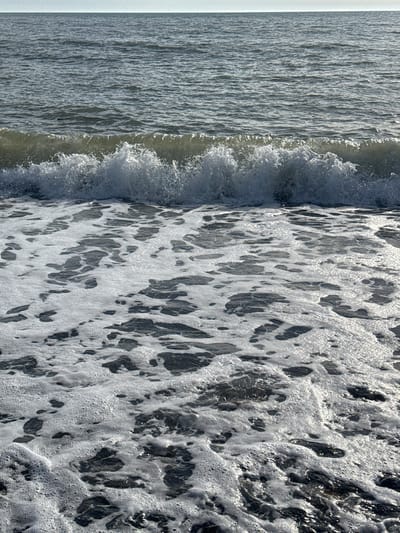 The image depicts a body of water, presumably the sea, with a wave crashing along a shoreline. The water transitions from a relatively calm surface on the left to frothy, foamy water on the right as the wave breaks. The foam creates a mosaic pattern of dark and light areas. The image is taken at an angle, making the scene appear vertically oriented. Although the image is from Gulripshi, Abkhazia, no landmarks are visible to confirm the location. There are no people present.
