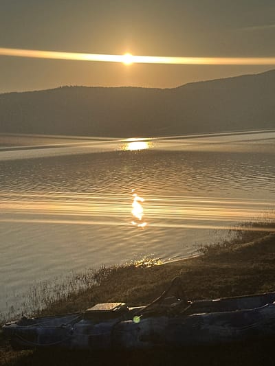 The image shows a serene lake scene at sunrise, likely near Batak, Bulgaria. The sun is low on the horizon, casting a warm, golden glow across the water and the surrounding hills. The lake's surface is rippled, reflecting the sunlight in shimmering patterns. On the shore, a camouflage-patterned kayak rests on the grassy bank. The foreground is dominated by the kayak and the edge of the water, with sparse vegetation. The background features rolling hills, silhouetted against the bright sky. The overall atmosphere is peaceful and tranquil.