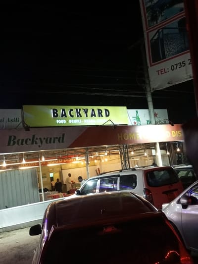 The image depicts a nighttime scene of a restaurant or bar named "Backyard" located in Kinanie ward, Kenya. The establishment features an illuminated sign with "BACKYARD" in prominent letters and "FOOD - DRINKS - NYAMA CHOMA" below, alongside Heineken branding. A lower sign states "Backyard HOME OF GOOD DISH."

The "Backyard" area appears to be a semi-open or open-air dining space, brightly lit by numerous suspended string lights. Visible within this area are at least three male individuals. One man, wearing a white coat or apron, stands near a counter or service area, facing towards the right. Another man in dark clothing is visible further to the left. A third man is partially obscured behind the white-coated individual. The interior includes tables and chairs, and a red fire extinguisher is mounted on a pillar. A corrugated metal wall forms part of the left boundary of the dining area.

In the immediate foreground, several vehicles are parked, partially obscuring the lower portion of the establishment. A dark-colored sedan (possibly maroon or dark red) is prominent in the center, viewed from its rear. Behind it, a white multi-purpose vehicle or small van is parked, with its side and rear visible, showing a partially obscured Kenyan license plate (appearing to begin with "KCR"). A light-colored sedan is partially visible on the far right.

Above the "Backyard" sign, the night sky is dark, with overhead electrical or communication wires visible. To the upper right, a vertical banner-style sign is partially visible on a pole, displaying images of decorative metalwork and a contact number "TEL: 0735 2XXXXXX", consistent with a Kenyan mobile number prefix. The overall scene suggests an active business operating during evening hours.