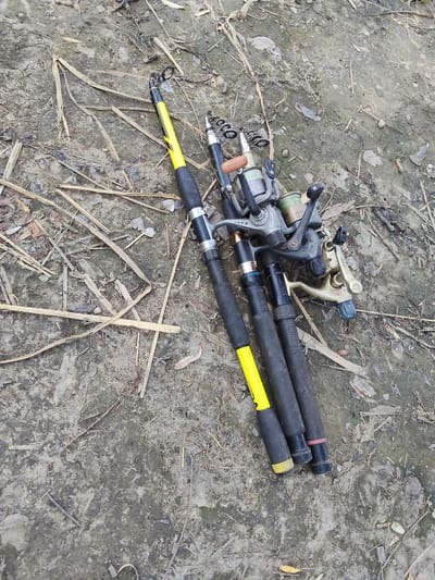 The image shows three fishing rods with reels lying on the ground. The ground appears to be dirt mixed with dried leaves and twigs, suggesting an outdoor setting. Two of the fishing rods have yellow accents on their handles, while the third has black handles. The reels are of various sizes and colors, with visible fishing line on at least one of them. The fishing rods are positioned diagonally across the frame, with the reels closer to the bottom right corner and the rod tips extending towards the upper left. There are no people visible in the image, and the scene is static, with the fishing rods simply resting on the ground. The lighting suggests it is daytime. Text visible on one of the reels reads "LONG CAST" and "340". The location context provided is Bilyaivka, Ukraine.