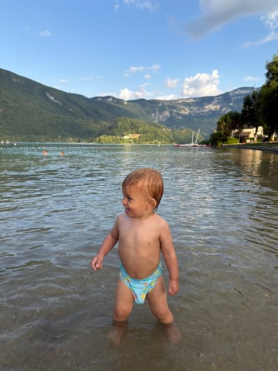 A toddler stands in shallow, clear water, facing to the right and looking down with a slight smile. The child has fair hair and is wearing blue swim trunks with a cartoon print. Their left arm is bent, and their right foot is submerged in the water. The water's surface is rippled, with sunlight reflecting off it. The background shows a rocky, sandy lakebed beneath the water, with gentle waves disturbing the surface.
Further in the background, across the water, is a shoreline lined with trees and buildings. A bright blue sky with scattered white clouds forms the upper background. A long line of buoys marks an area in the water. The overall impression is a sunny day at a lake, likely a beach or swimming area. The scene suggests a relaxed, recreational atmosphere, with the toddler enjoying the water. The location is identified as Novalaise, France. There is no visible text in the image.