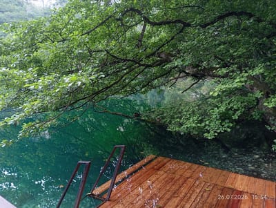 A wooden dock juts out into a clear, turquoise body of water, surrounded by lush green foliage. The dock is wet, with water droplets visible on its surface, suggesting recent rain. Metal railings are attached to the dock, leading down into the water. In the distance, a duck is perched on a fallen branch extending over the water. The water itself is transparent, allowing the rocky bottom to be seen in places. The background features a forested hillside and a hint of rock formations. A timestamp, "26.07.2026 15:34", is visible in the lower right corner of the image. This scene appears to be a serene natural setting, possibly a lake or river in Verkhnyaya Balkariya, Russia.